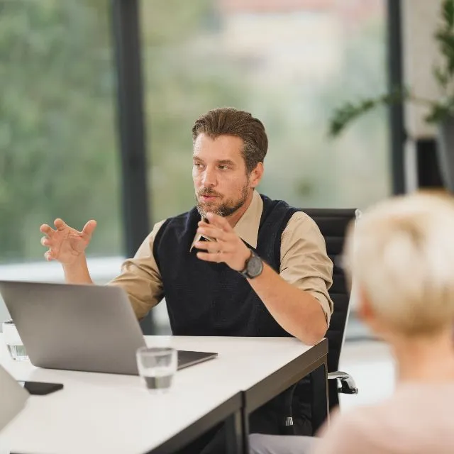 A manager talks with two people around a desk, laptop open